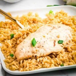 Side shot of a piece of chicken with rice garnished with green onions on a square white plate with a gold fork on the side.