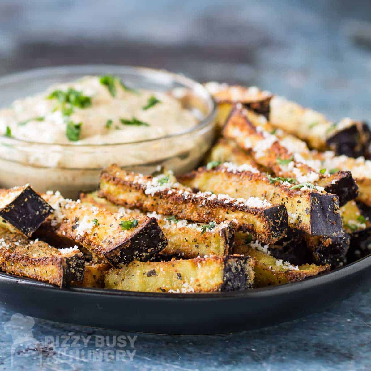 Side close up shot of eggplant fries garnished with basil and parmesan on a black plate with a clear bowl of chipotle basil dip garnished with more fresh basil.
