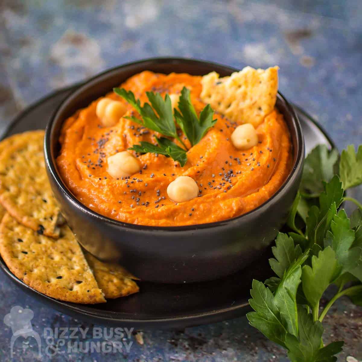 Side angled shot of red pepper hummus in a black bowl on a black plate garnished with pepper, chickpeas, and herbs, and crackers and more herbs on the side.