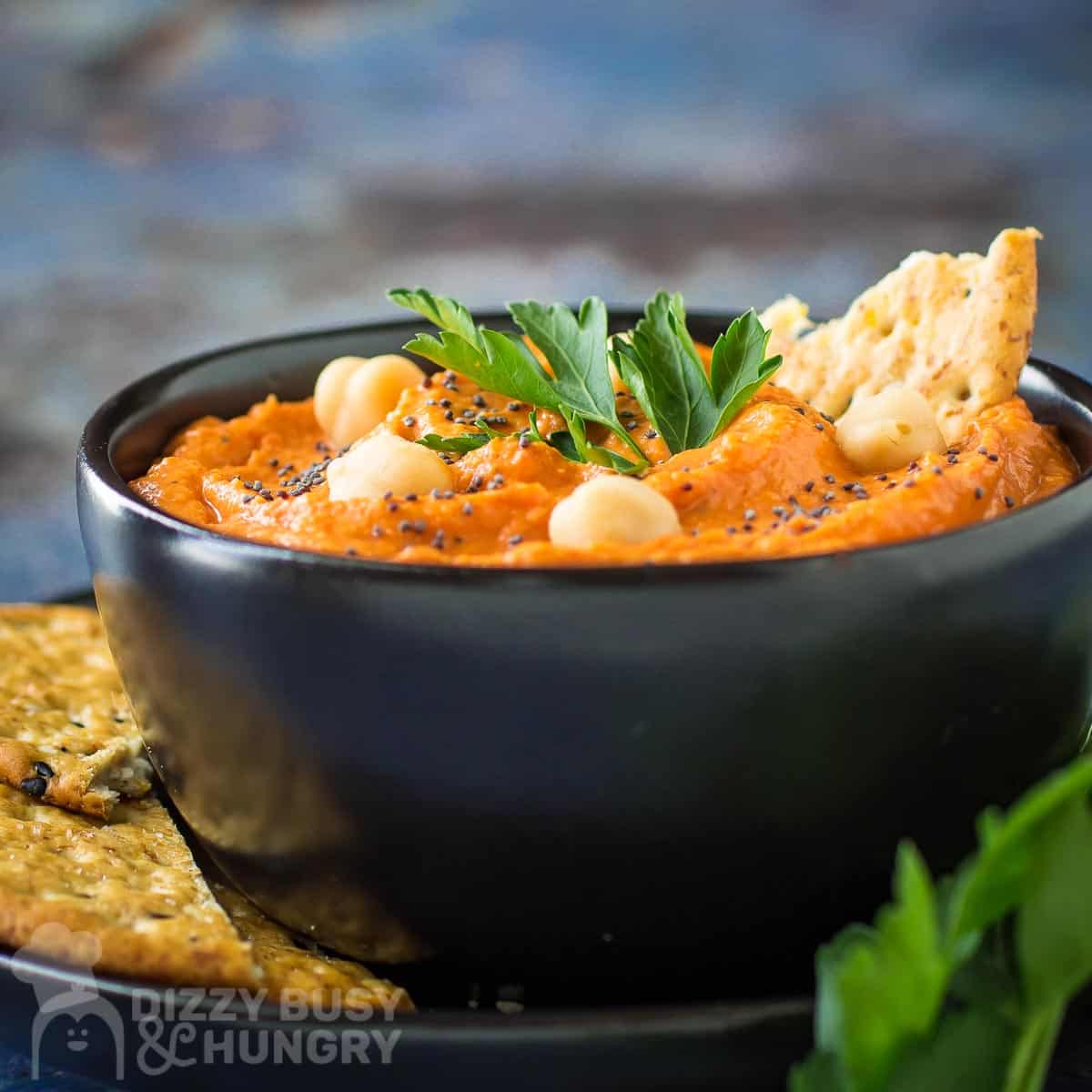 Side close up shot of red pepper hummus in a black bowl on a black plate garnished with pepper, chickpeas, and herbs, and crackers on the side.