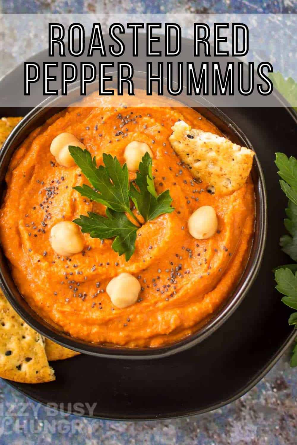 Overhead shot of red pepper hummus in a black bowl on a black plate garnished with pepper, chickpeas, and herbs, and crackers on the side.