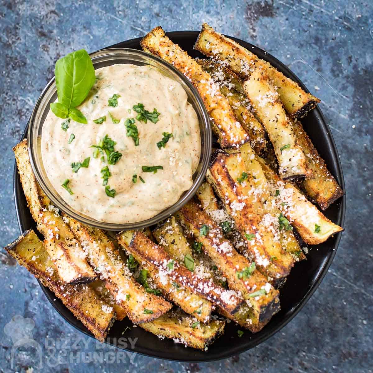 Overhead shot of eggplant fries garnished with basil and parmesan on a black plate with a clear bowl of chipotle basil dip garnished with more fresh basil.