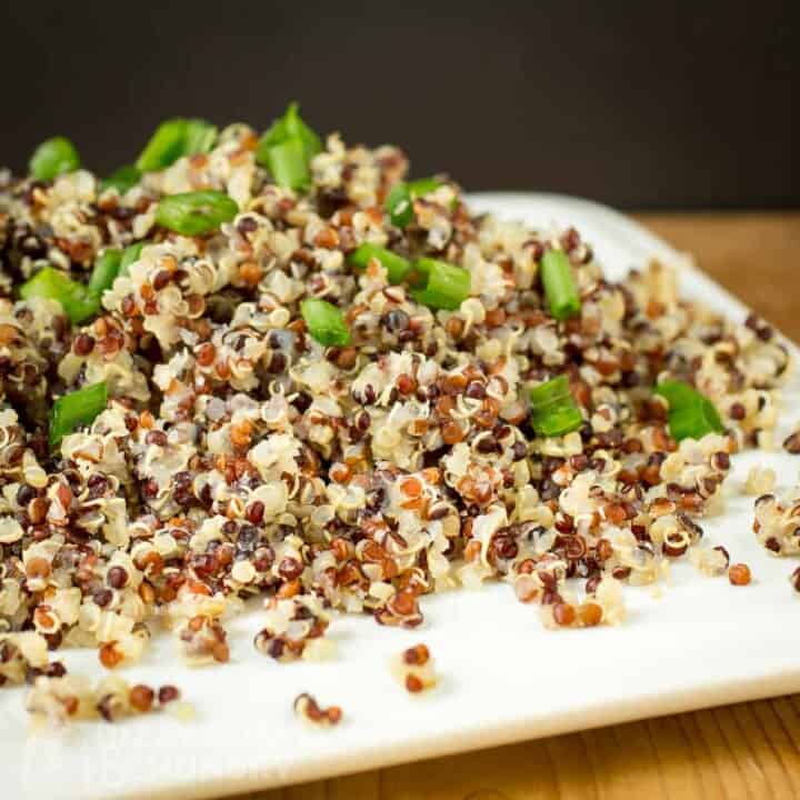 Side shot of quinoa garnished with green onions on a square white plate on a wood table.