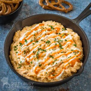 Side angled shot of buffalo chicken dip garnished with buffalo sauce, sour cream, and herbs in a cast iron skillet with a bowl of pretzels in the background.