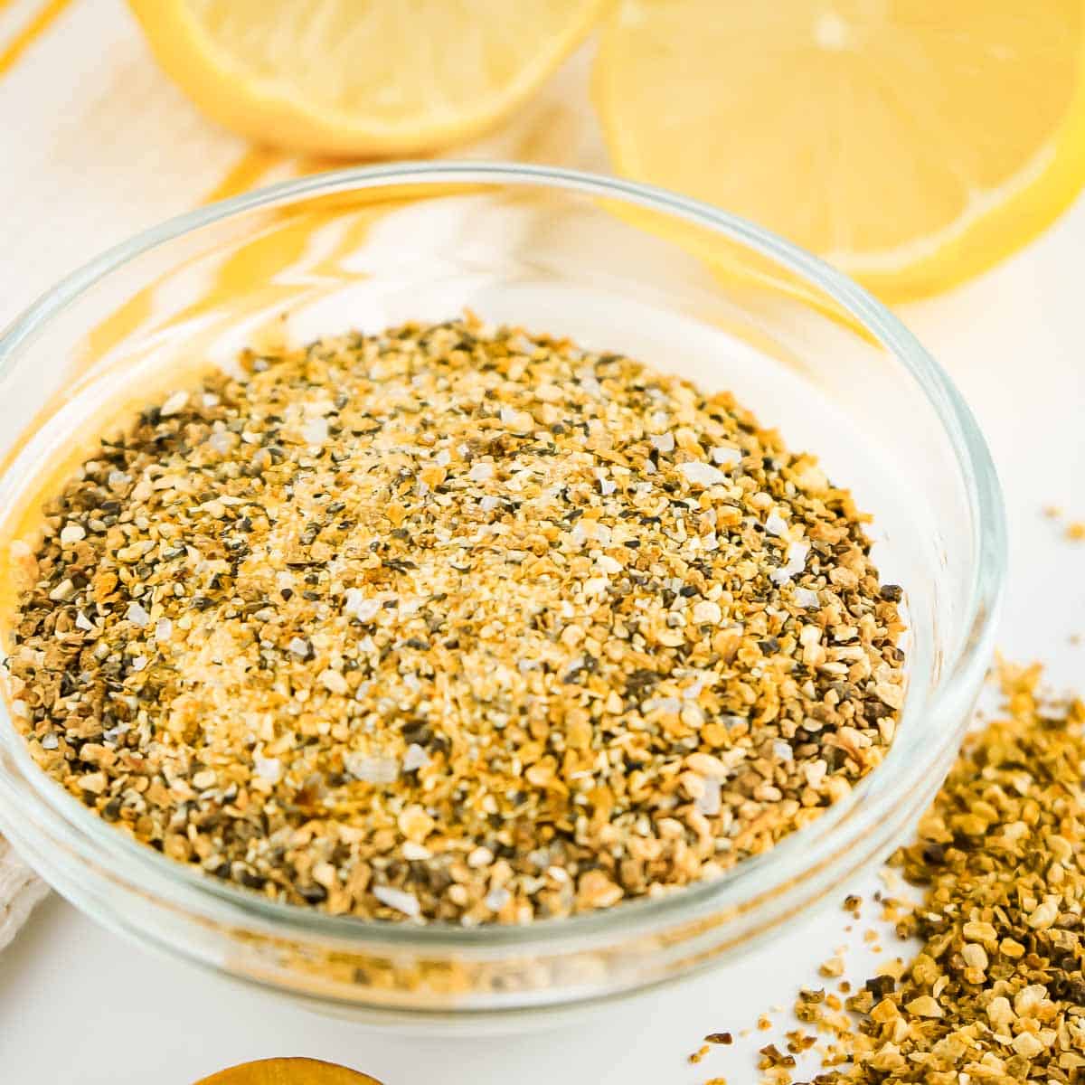 Side angled shot of seasoning in a small clear bowl on a white surface with two halved lemons in the background.