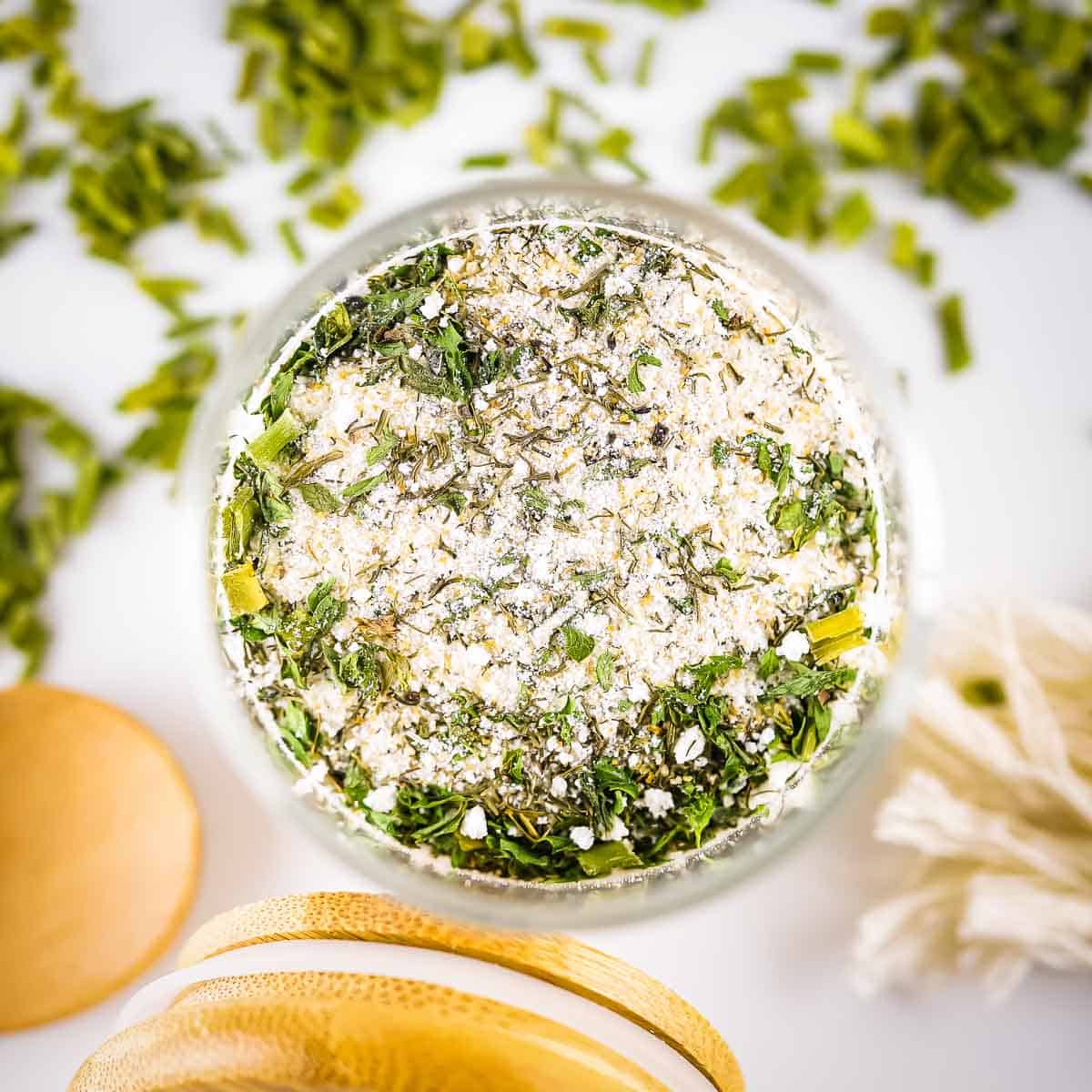 Overhead shot of a glass jar full of ranch seasoning with the wooden lid leaning on the side all on a white countertop with herbs scattered around.