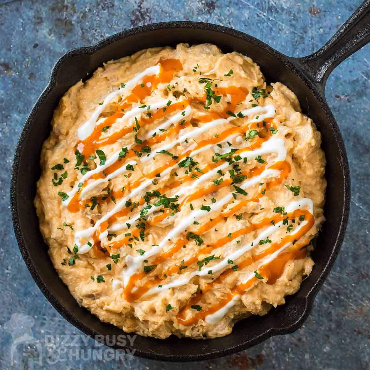 Overhead shot of buffalo chicken dip garnished with buffalo sauce, sour cream, and herbs in a cast iron skillet on a blue surface.