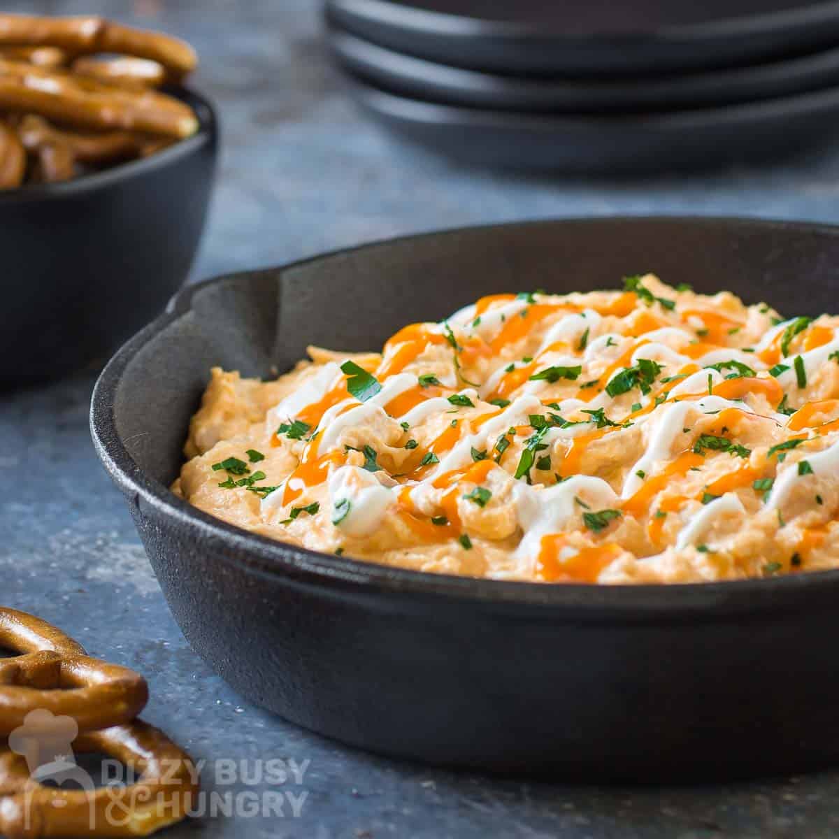 Side angled shot of buffalo chicken dip garnished with buffalo sauce, sour cream, and herbs in a cast iron skillet with a bowl of pretzels and a stack of plates in the background.