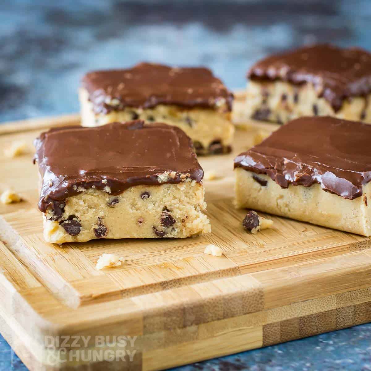 Side shot of multiple cookie dough bars cut in squares on a wooden cutting board on a blue surface.