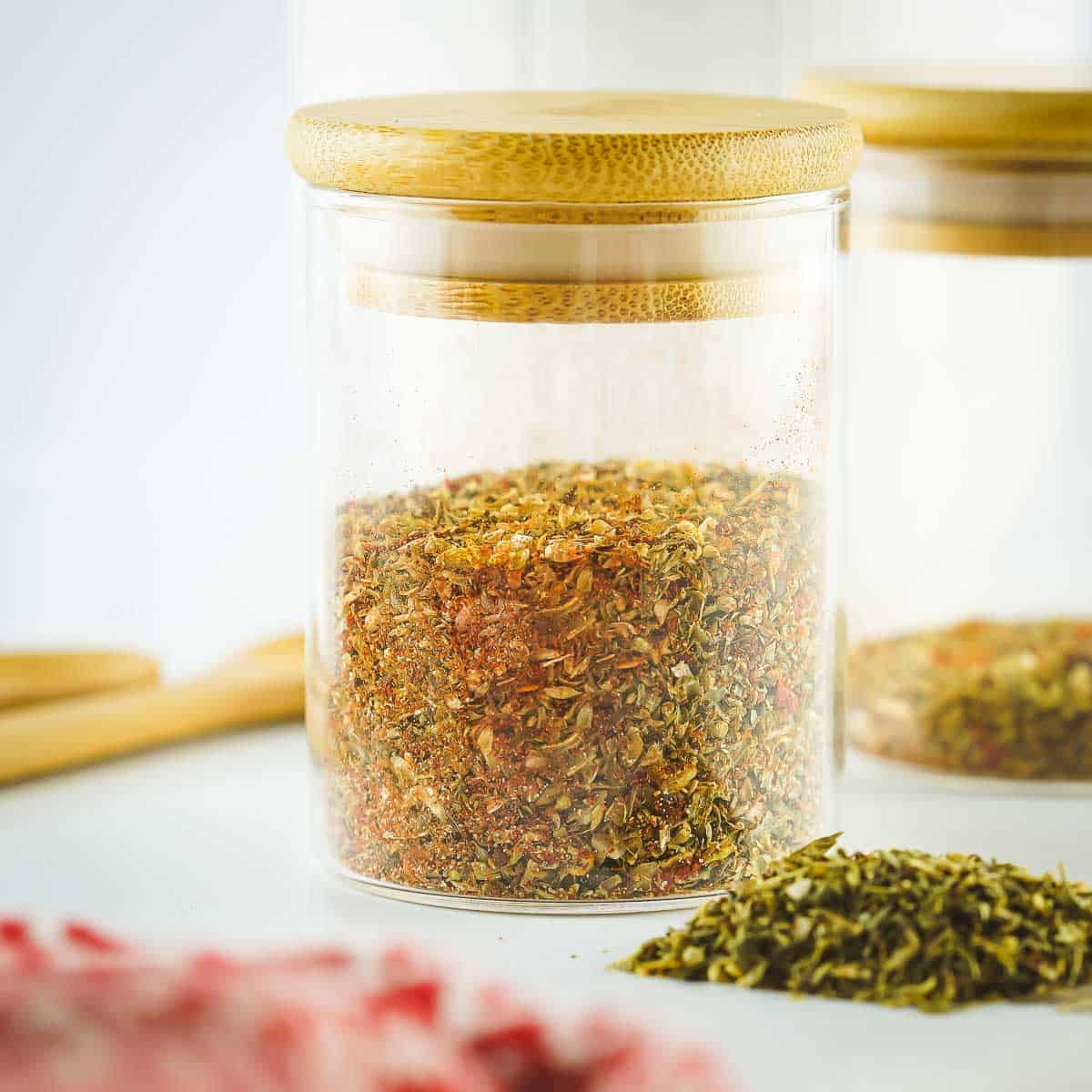 Side shot of pizza spices in an glass jar with a wooden cap on a white surface with fresh herbs and a wooden spoon on the side.