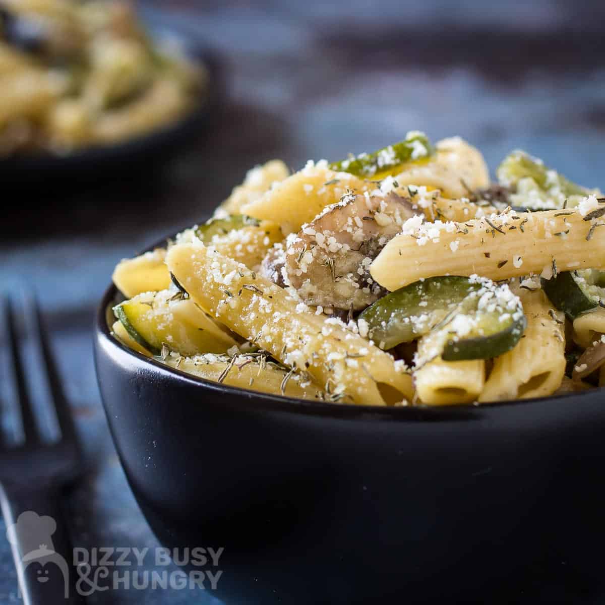 Side close up shot of mushroom zucchini pasta in black bowl with a black fork on the side with a blue background.