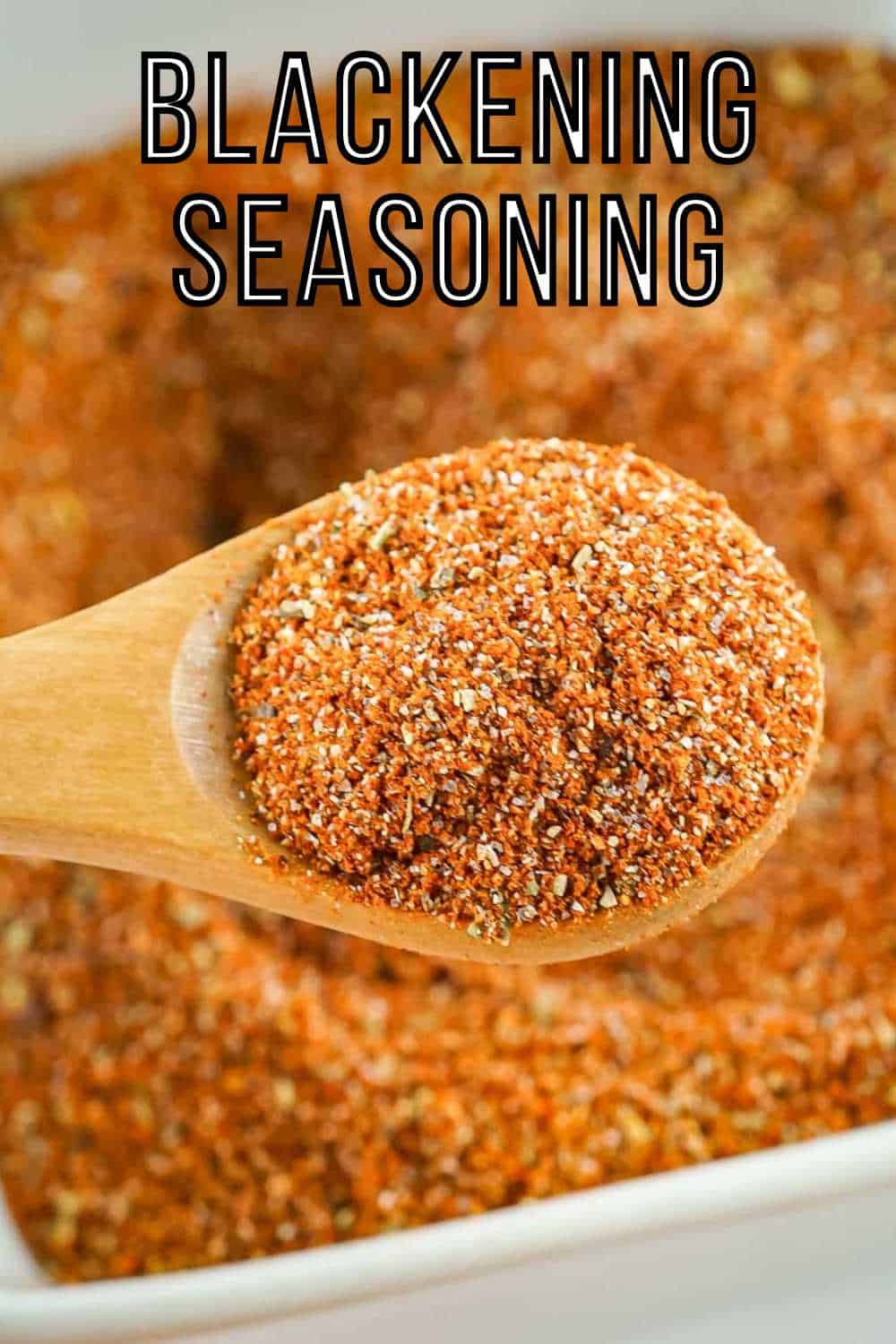 Overhead close up shot of a wooden spoon holding a scoop of seasoning over a white bowl full of blackened seasoning on a white surface.