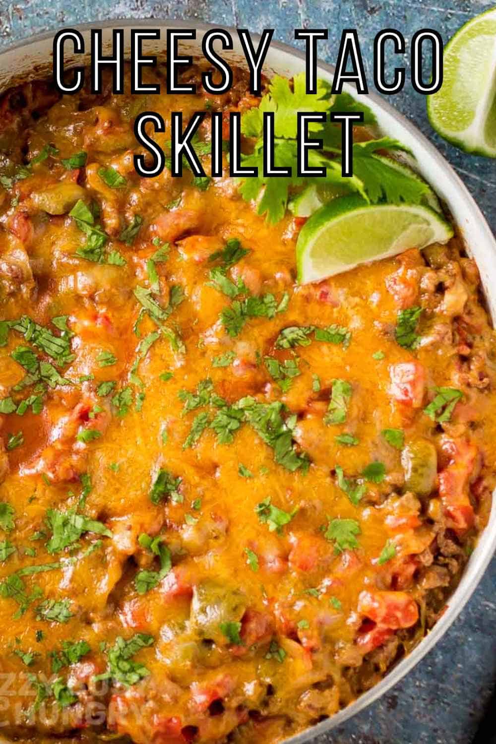 Overhead shot of cheesy taco meat garnished with fresh parsley and lime wedges in a black and white skillet on a whicker placemat.