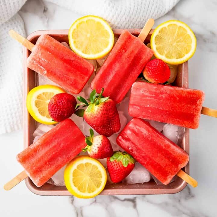 Overhead shot of popsicles on a bed of ice in a pink bowl with lemon slices and strawberries.