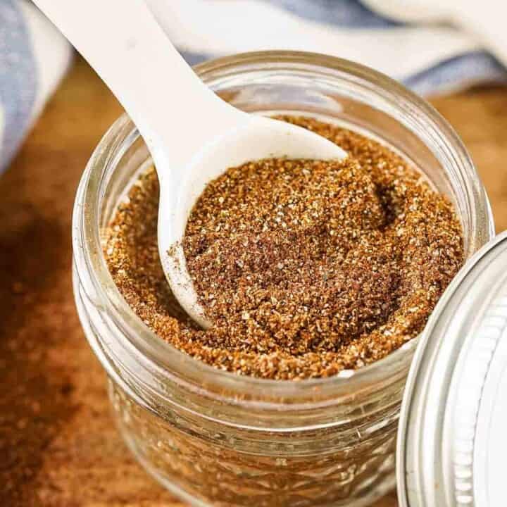 Side close up shot of a white spoon scooping some chili seasoning out of a clear mason jar full of seasoning on a wooden table.