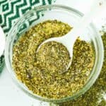 Overhead shot of greek spice mix in a clear jar with a white spoon inside on a white surface with more spice mix on the side.