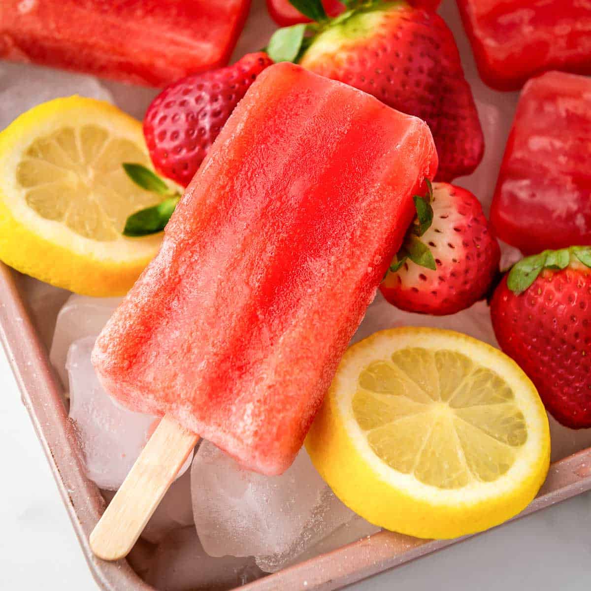 Angled close up shot of popsicles on a bed of ice in a pink bowl with lemon slices and strawberries.