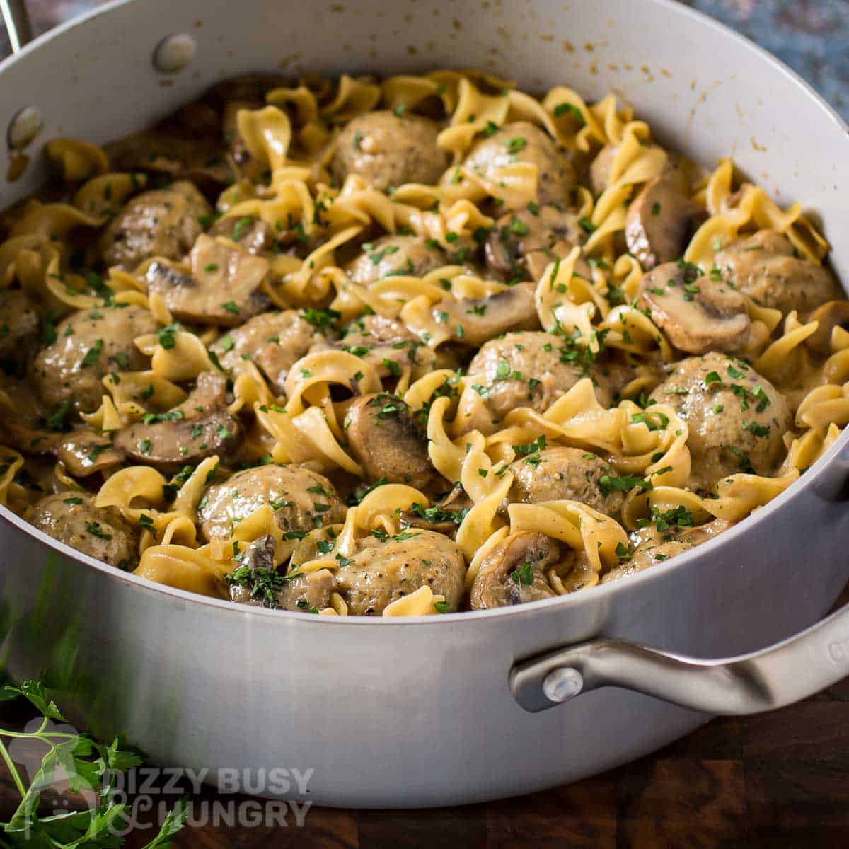 Side angled shot of a large silver pan full of meatball stroganoff garnished with fresh herbs on a wooden surface.