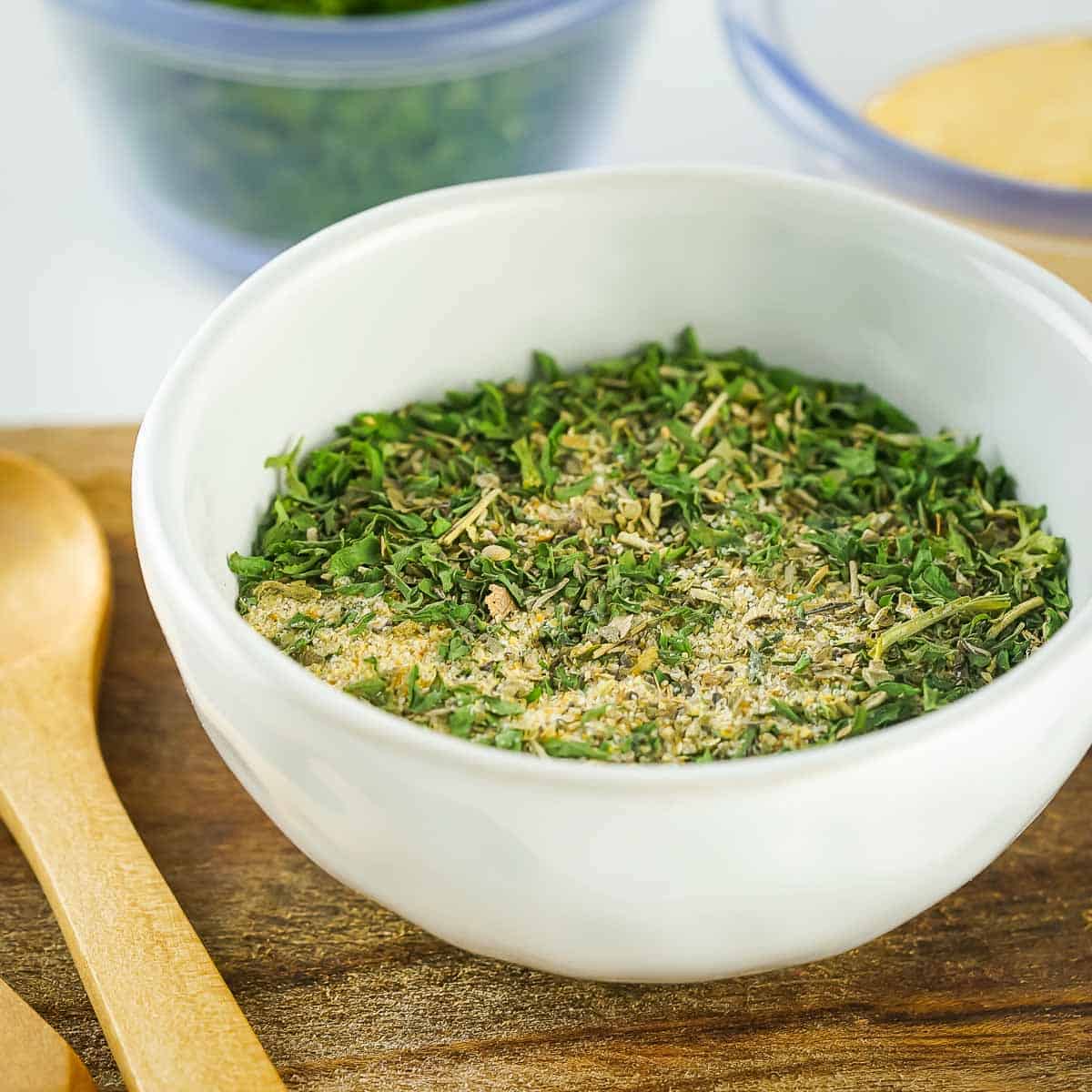 Side angled shot of seasoning in a white bowl on a wooden cutting board with a wooden spoon on the side.