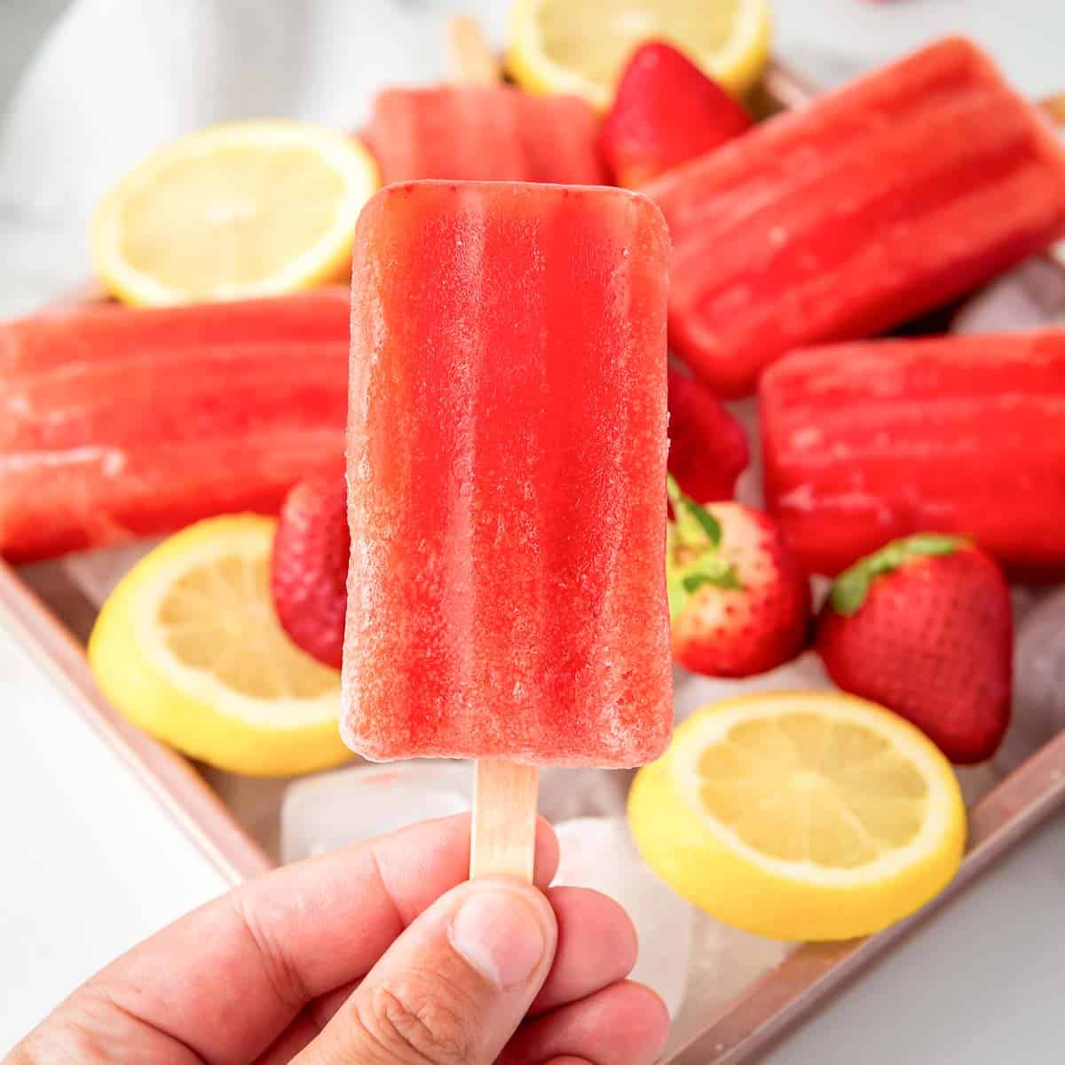 Side shot of a hand holding up one popsicle in front of a tray of popsicles laying on a bed of ice, lemon slices, and strawberries.