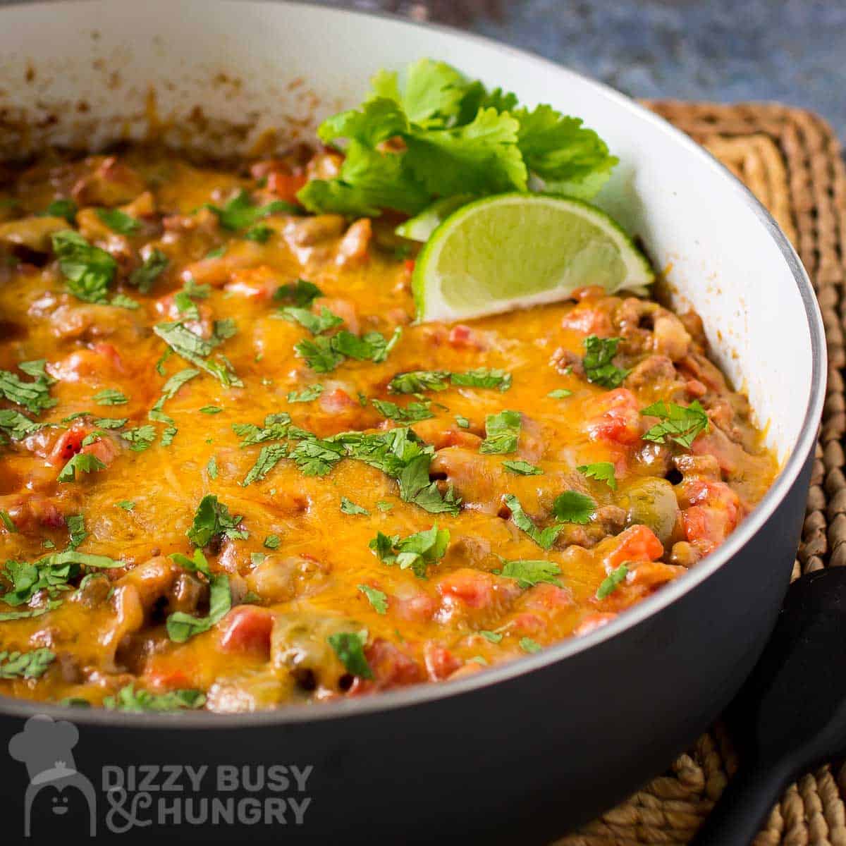 Side close up shot of cheesy taco meat garnished with fresh parsley and lime wedges in a black and white skillet on a whicker placemat.