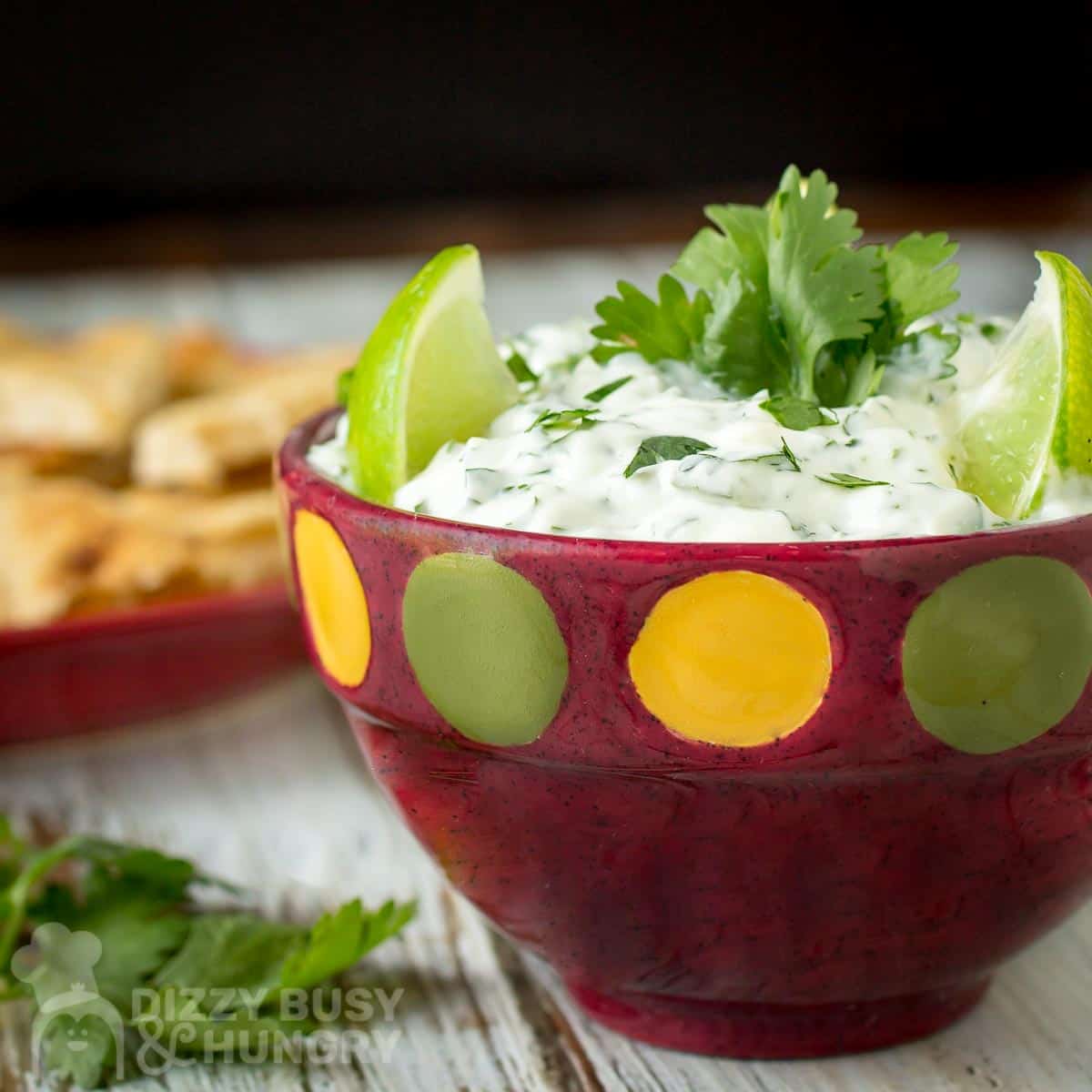 Close up shot of cilantro lime dip garnished with limes and cilantro in a red polka dot bowl with chips in the background.