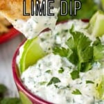 Close up shot of a chip being dipped into cilantro lime dip with sliced limes and cilantro in the background.