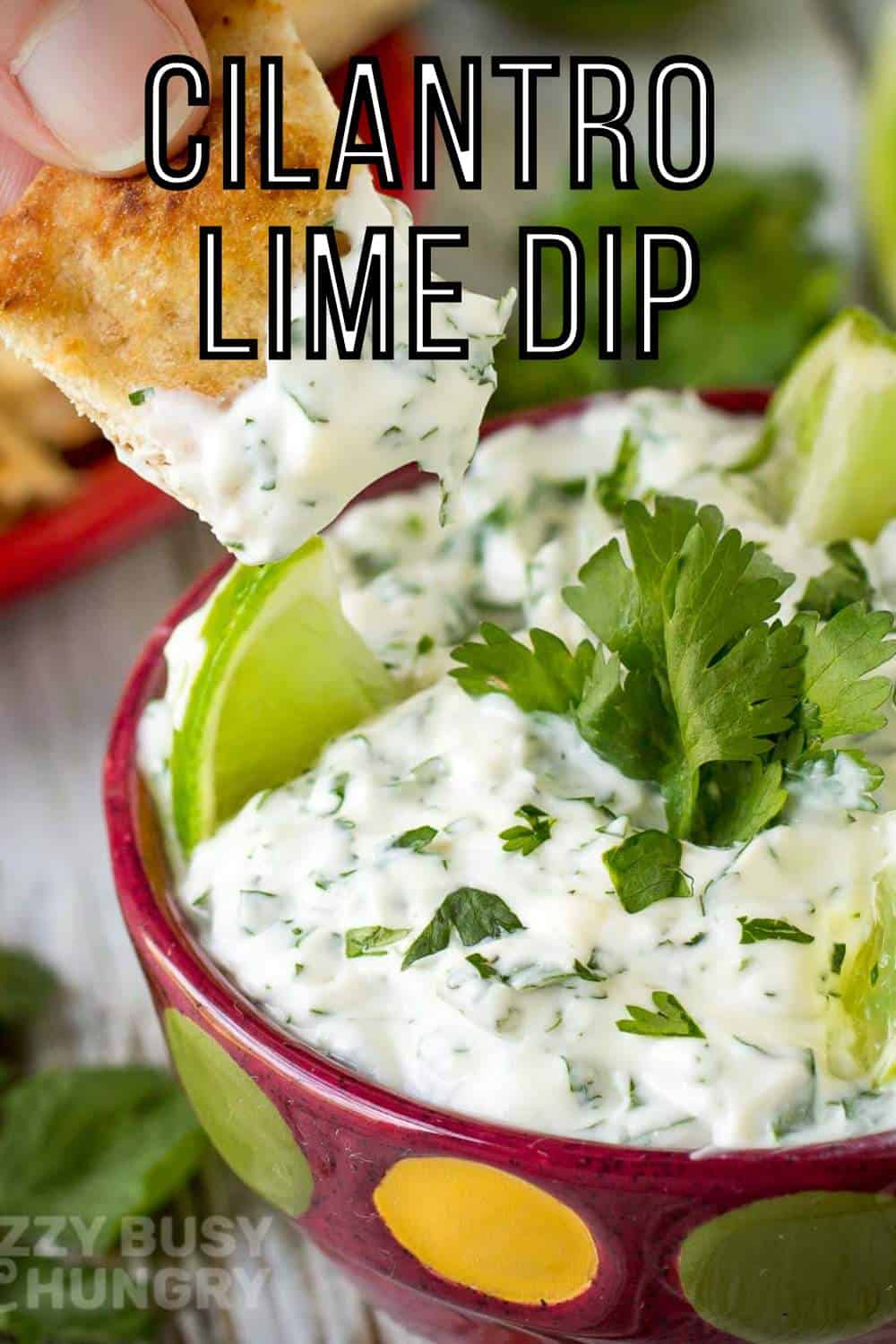 Close up shot of a chip being dipped into cilantro lime dip with sliced limes and cilantro in the background.