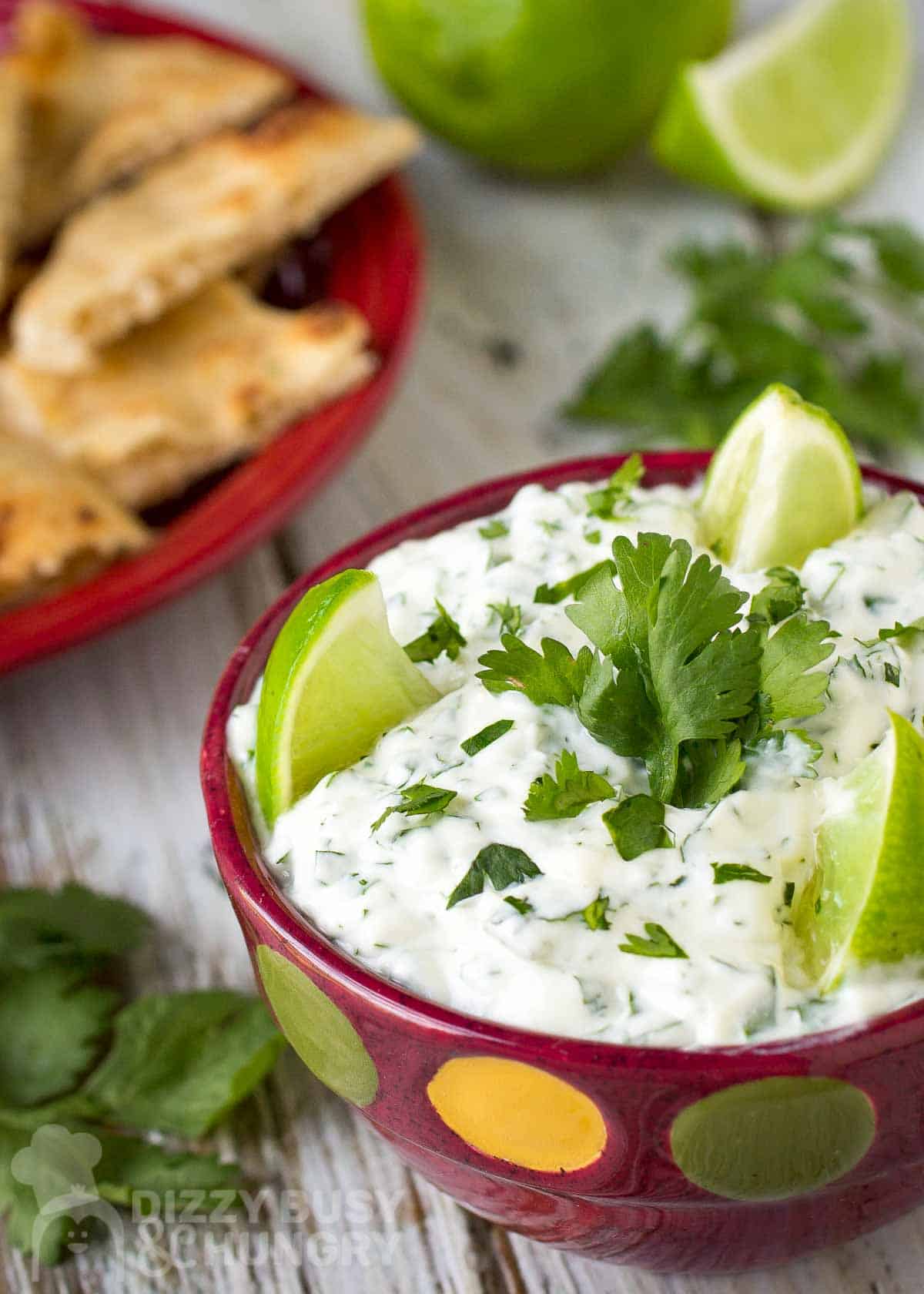 Close up angled shot of cilantro lime dip garnished with limes and cilantro in a red polka dot bowl with chips in the background.