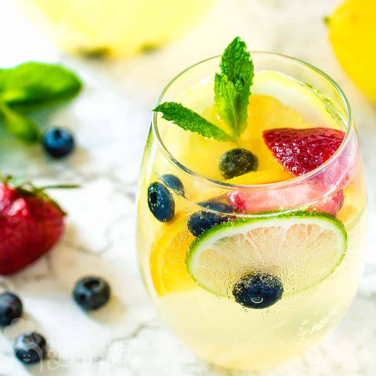 Side shot of a glass wine tumbler with sangria garnished with mint and fruits on a marble surface with a pitcher of it in the background and fruits on the side.