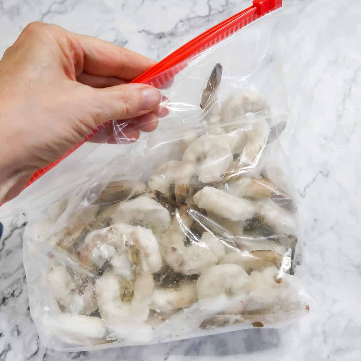 Overhead shot of a hand zipping up a clear ziplock bag full of frozen shrimp on a white marble surface.