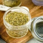 Side shot of a small mason jar with seasoning on a wooden cutting board with the lids leaning on the side and a white measuring spoon in the background.