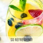 Close up shot of a glass wine tumbler with sangria garnished with mint and fruits on a marble surface with a pitcher of it in the background.