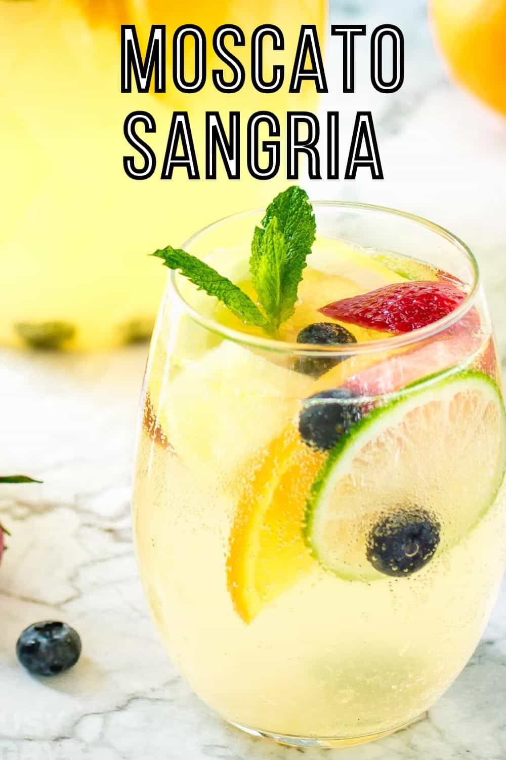 Side shot of a glass wine tumbler with sangria garnished with mint and fruits on a marble surface with a pitcher of it in the background.