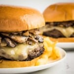Side close up shot of two white plates each with a mushroom burger and French fries on the side on a white surface.