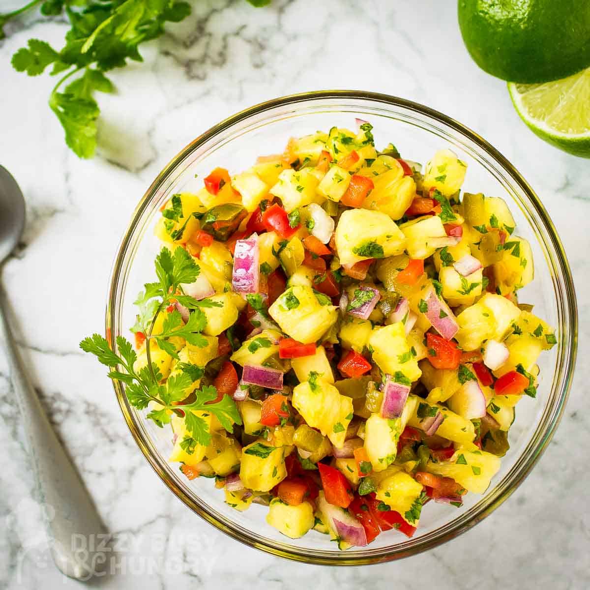 Overhead shot of pineapple salsa garnished with cilantro in a clear bowl on a white surface with more herbs and halved limes in the background.