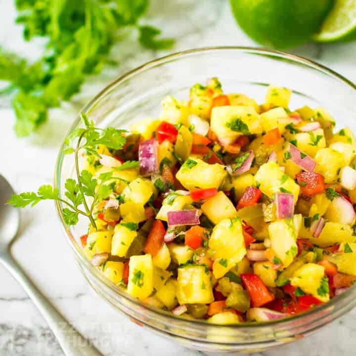 Side shot of pineapple salsa garnished with cilantro in a clear bowl on a white surface with more herbs and halved limes in the background.
