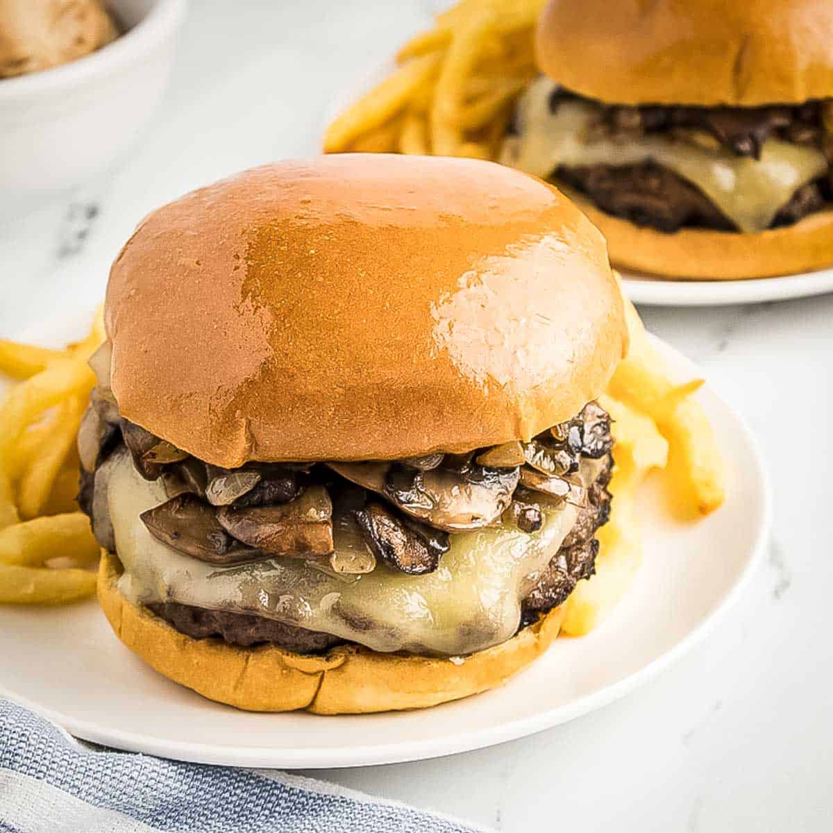 Side shot of two white plates each with a mushroom burger and French fries on the side with a blue and white striped cloth on the side.