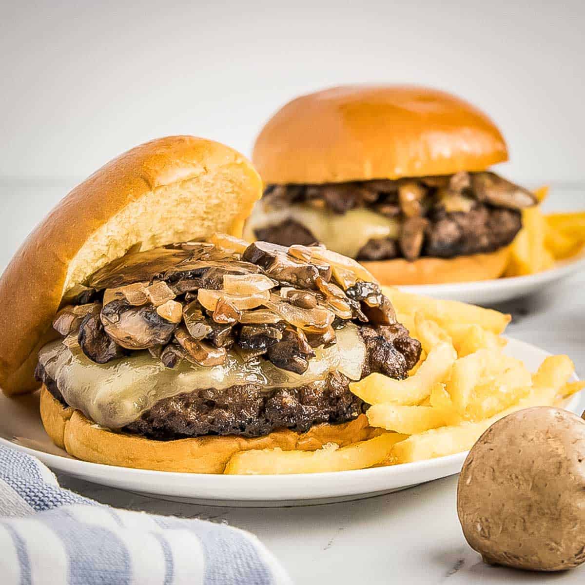 Side shot of two white plates each with a mushroom burger and French fries on the side with a mushroom and blue and white striped cloth on the side.