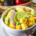 Close up side shot of mango avocado salad in a white bowl with a gold spoon on a white plate.