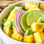 Side shot of mango avocado salad in a white bowl on a white plate with a wooden bowl with fruit in the background.