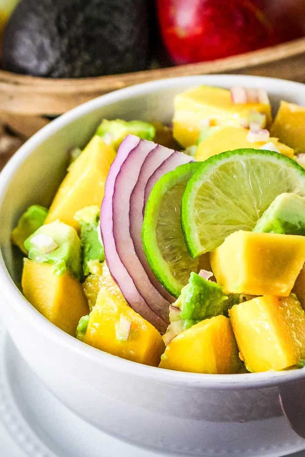 Side shot of mango avocado salad in a white bowl on a white plate with a wooden bowl with fruit in the background.