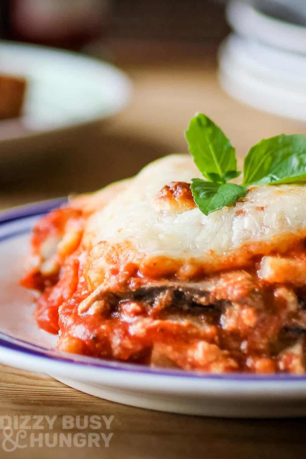 Side shot of eggplant polenta lasagna garnished with basil on a white and blue plate on a wooden table.