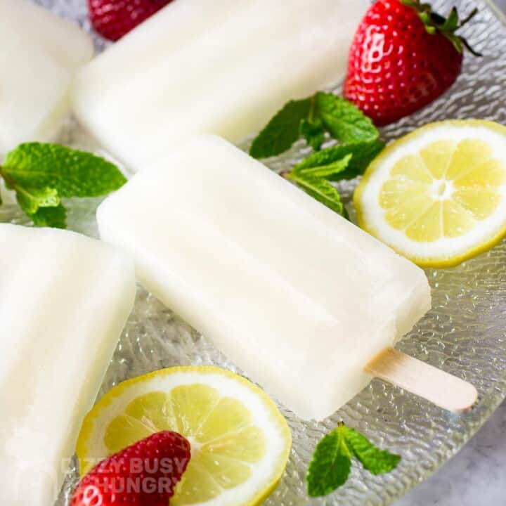 Side shot of lemonade popsicles on a glass plate with mint, lemon slices, and strawberries scattered around.
