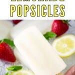 Side shot of a hand holding up a lemonade popsicle over a glass plate with popsicles, strawberries, lemon slices, and mint.