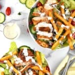 Overhead shot of two white plates with Pittsburgh salad drizzled with ranch with a small glass jar of ranch, tomatoes, and cucumbers on the side on a white surface.