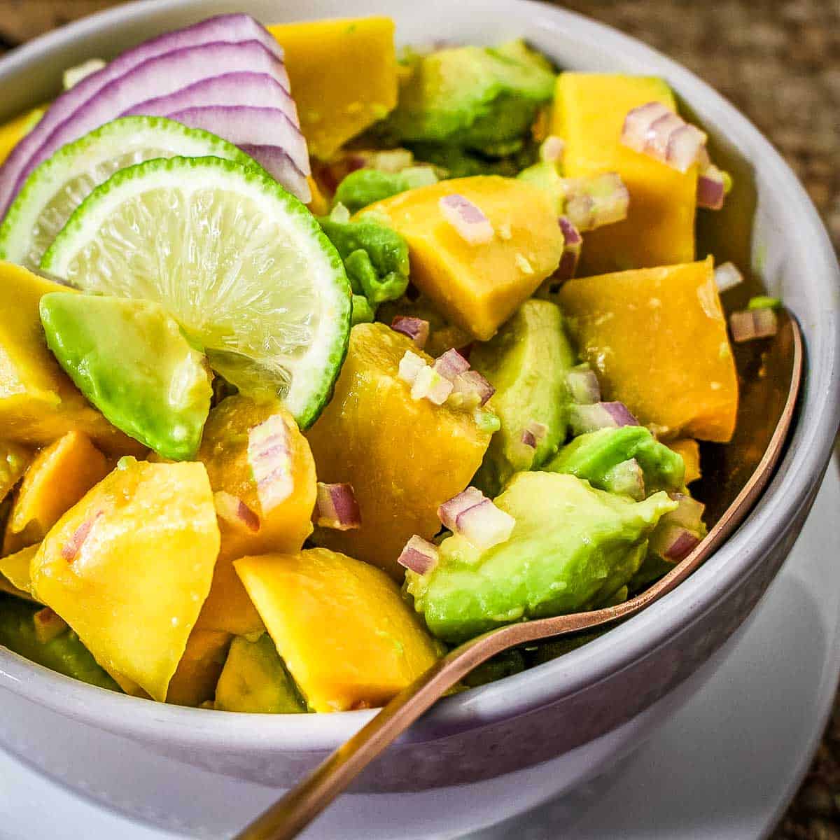 Close up shot of mango avocado salad in a white bowl with a gold spoon on a white plate.