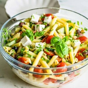 Side shot of pasta salad garnished with basil in a large clear bowl on a marble table.