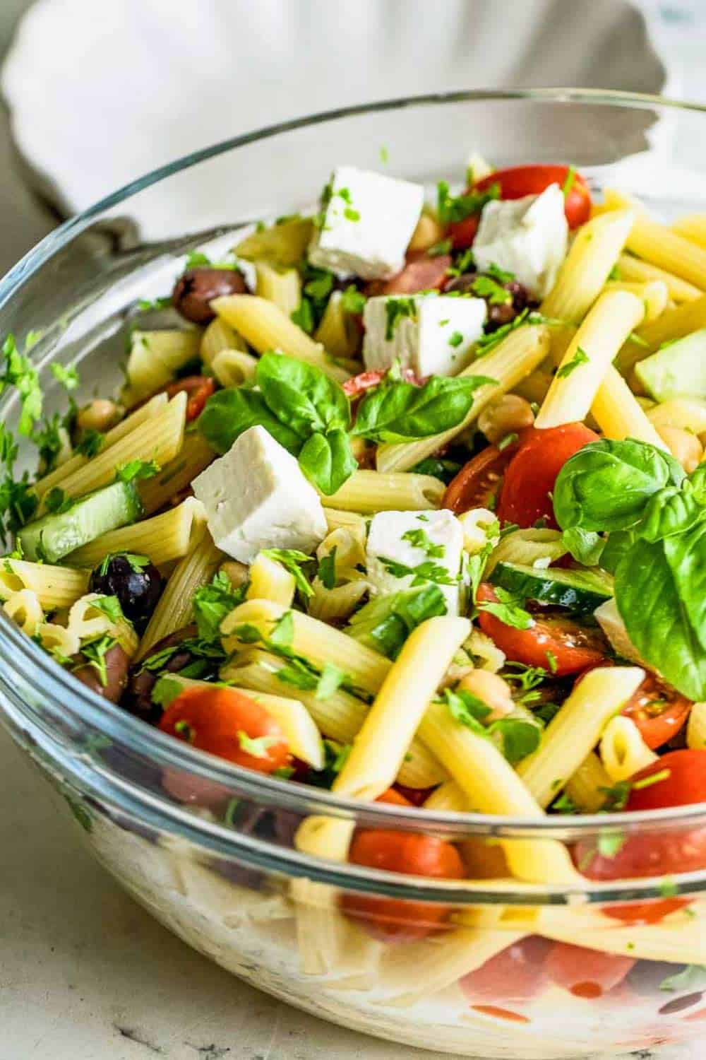 Close up shot of pasta salad garnished with basil in a large clear bowl on a marble table.