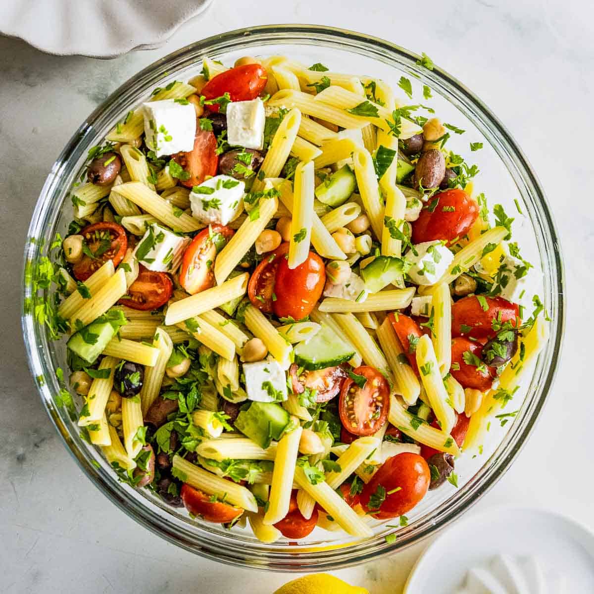 Overhead shot of pasta salad garnished with basil in a large clear bowl on a marble table.